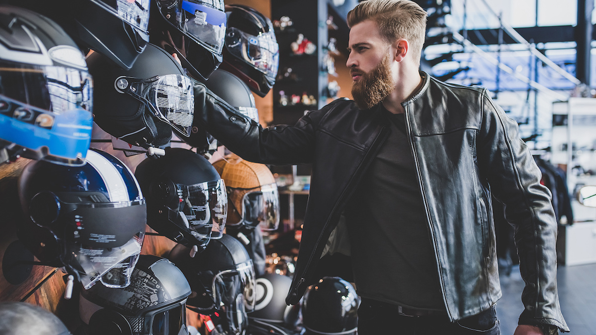 Joven barbudo en la tienda de motocicletas buscando comprar un nuevo casco de motocicleta.